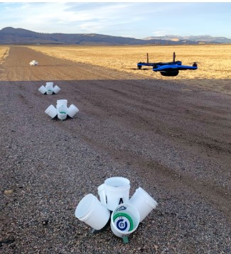 A drone hovers over a field with scattered planting containers on the ground, showcasing agricultural technology in action.